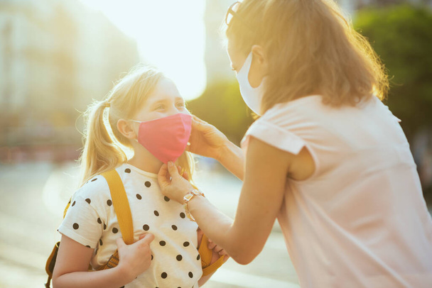 Life during covid-19 pandemic. young mother and school girl with masks and yellow backpack getting ready for school outside. - Фото, изображение