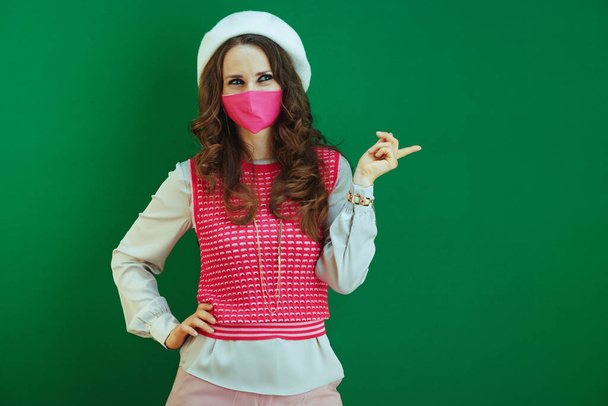 Life during coronavirus pandemic. pensive elegant female in pink sleeveless shirt with pink medical mask pointing at something isolated on green background. - 写真・画像