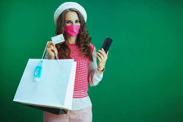 Life during coronavirus pandemic. young woman shopper in white beret with pink medical mask, shopping bags, sanitiser and credit card buying on internet on a smartphone against green background. - Фото, изображение