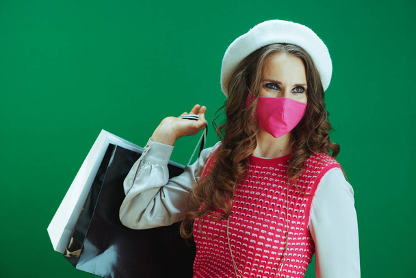 Life during coronavirus pandemic. trendy female in white beret with pink medical mask and shopping bags against green background. - Foto, imagen