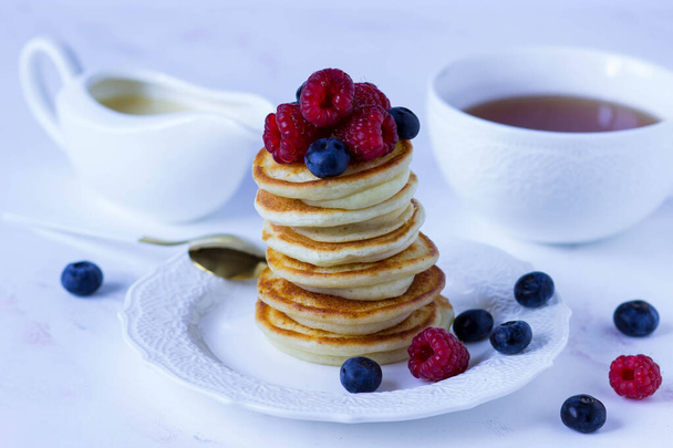 A stack of pancakes with raspberries and blueberries on a white plate against the background of a cup of tea and a gravy boat with condensed milk - Fotografie, Obrázek