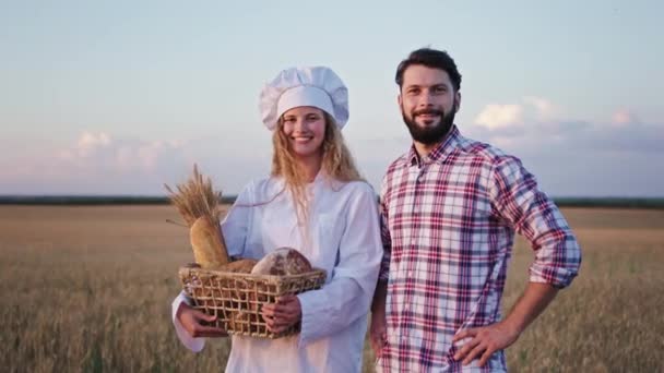 Portrait of a farmer man and baker woman in front of the camera standing together lady holding a basket full of fresh baked bread in the middle of the wheat field - Materiaali, video