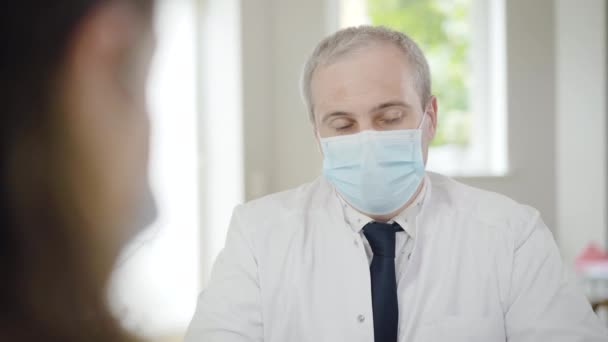 Mid-adult doctor in face mask consulting patient on Covid-19 pandemic. Blurred patient at front shaking head. Portrait of professional Caucasian specialist talking to woman in hospital. - Filmati, video