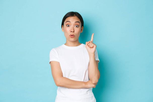 Lifestyle, beauty and shopping concept. Excited asian girl in white t-shirt, raising index finger, have suggestion, think-up plan or solution, saying plan, standing blue background - Fotografie, Obrázek