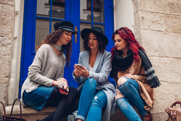 Three young women friends looking at smartphone on city street. Stylish girls talking and having fun. Group of girlfriends spend time together - Photo, Image