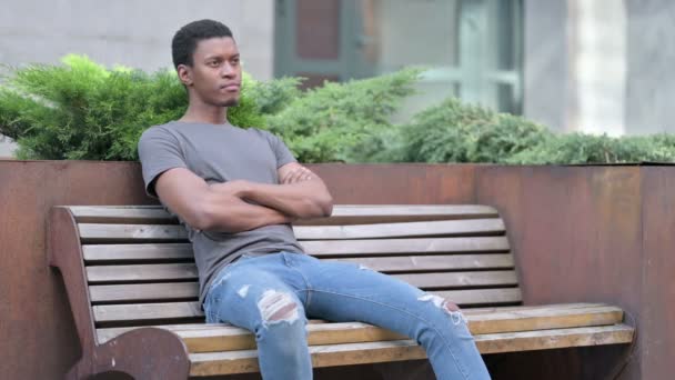 Pensativo joven africano joven sentado en el banco y pensando  - Imágenes, Vídeo