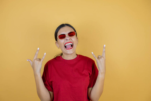 Young beautiful woman wearing red sunglasses over isolated yellow background shouting with crazy expression doing rock symbol with hands up.Young beautiful woman wearing red sunglasses over isolated yellow background shouting with crazy expression do - Photo, Image