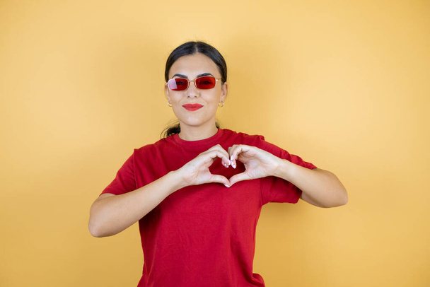Jeune belle femme portant des lunettes de soleil rouges sur fond jaune isolé souriant en amour montrant le symbole du cœur et la forme avec les mains. - Photo, image