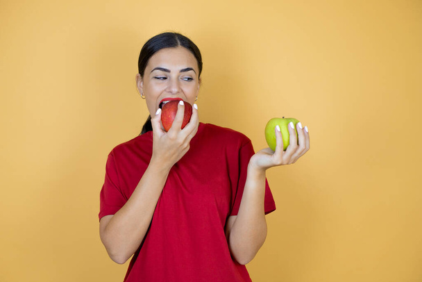 Joven hermosa mujer con camiseta roja de pie sobre fondo amarillo insolado elegir entre manzana roja y manzana verde. - Foto, Imagen