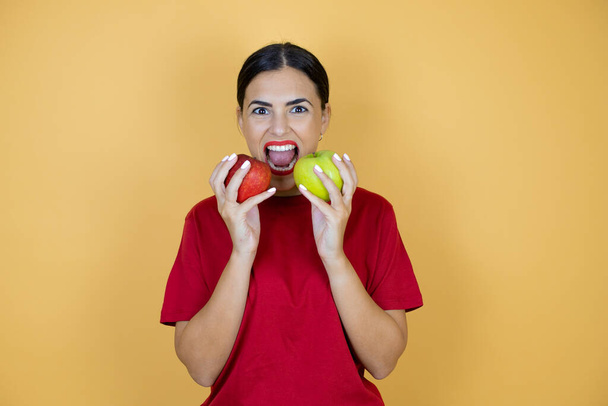Młoda piękna kobieta w czerwonej koszulce stojąca nad osamotnionym żółtym tłem jedząca czerwone jabłko i zielone jabłko. - Zdjęcie, obraz