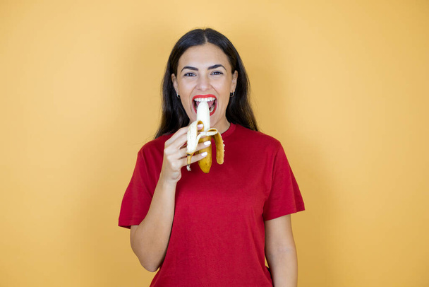 Junge schöne Frau trägt legeres rotes T-Shirt auf gelbem Hintergrund und isst eine Banane. - Foto, Bild