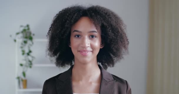 Kıvırcık saçlı genç Amerikalı kadın odasında kameranın önünde duruyor, beyaz dişleriyle geniş bir şekilde gülümsüyor, neşeli bir şekilde görünüyor, çekicilik kavramı, açık kalp - Video, Çekim