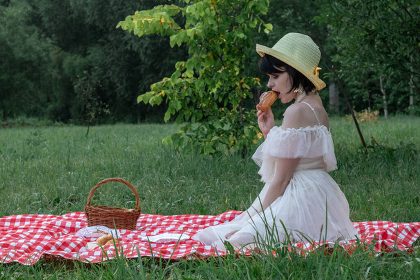 Όμορφη ρομαντική νεαρή γυναίκα με σκούρα κοντά μαλλιά σε ένα ψάθινο καπέλο και λευκό φόρεμα κάθεται σε ένα πικνίκ, στη φύση - Φωτογραφία, εικόνα