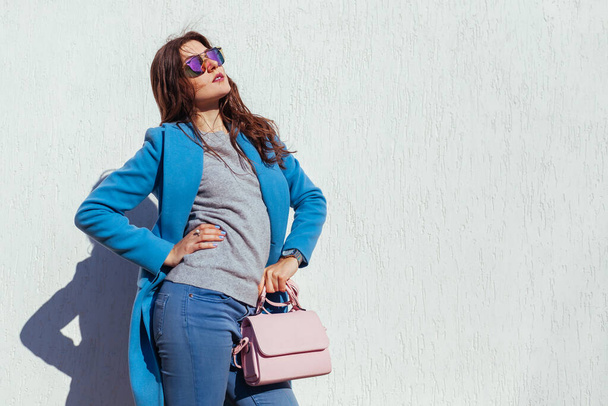 Young woman holding stylish purse and wearing trendy blue coat, sunglasses. Autumn female clothes and accessories. Fashion - Photo, Image