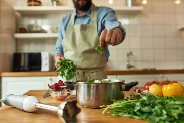 Cropped shot of man, chef cook adding basil leaf to the pot with chopped vegetables while preparing healthy meal, soup in the kitchen. Cooking at home, Italian cuisine concept - Fotografie, Obrázek