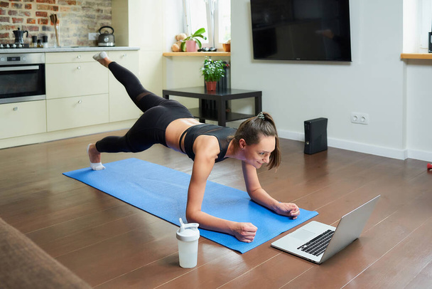 Una chica deportiva en un traje ajustado de entrenamiento está haciendo ejercicios en las nalgas con un video en línea en un ordenador portátil. Una entrenadora dirigiendo una clase de fitness remota en la alfombra azul de casa. - Foto, imagen