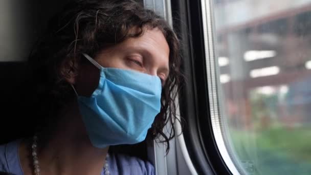 Zakenvrouw met vermoeide ogen gaat op een treinreis kijkend uit het raam. Jonge vrouw in een wegwerp medisch masker kijken naar lopende gebouwen, arme buurten, coronavirus vernietigt de economie - Video