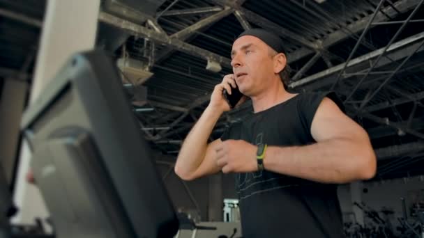 Hombre atleta está hablando en un teléfono celular mientras se ejecuta en una cinta de correr - Metraje, vídeo