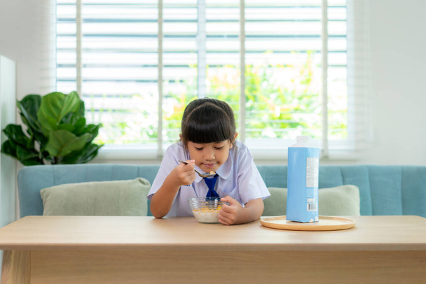 Asiatique élémentaire écolière fille en uniforme manger des céréales petit déjeuner avec du lait dans la routine de l'école du matin pour la journée dans la vie se préparer pour l'école. - Photo, image