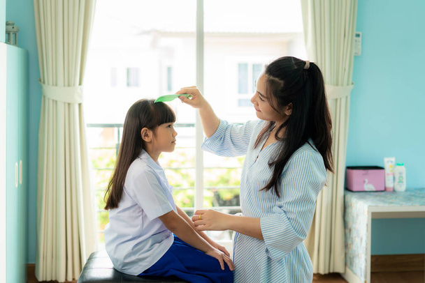 Ασιάτισσα μητέρα χτενίζει τα μαλλιά της κόρης της το πρωί πριν πάει σχολείο στο σαλόνι στο σπίτι. Η πρωινή σχολική ρουτίνα για την ημέρα της ζωής ετοιμάζεται για το σχολείο - Φωτογραφία, εικόνα