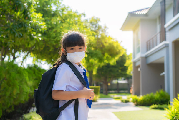 Πορτρέτο του χαριτωμένο κορίτσι της Ασίας δημοτικό σχολείο στην ιατρική μάσκα μπροστά από το σπίτι κατά τη διάρκεια COVID-19 πανδημία. Η πρωινή σχολική ρουτίνα για την ημέρα στη ζωή ετοιμάζεται για schoo - Φωτογραφία, εικόνα