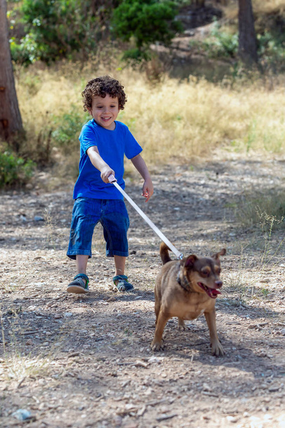 Kind met zijn kleine hondje loopt aan de lijn in een natuurpark lachend terwijl de hond zich uitstrekt en de riem aandraait - Foto, afbeelding