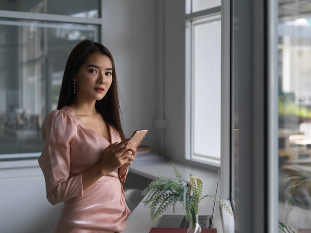 Young businesswoman holding smartphone while looking at the camera in office room  - Valokuva, kuva