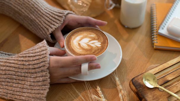 Oříznutý záběr mladé ženy držící latte art kávu na dřevěném stole s papírnictví  - Fotografie, Obrázek