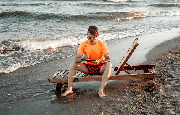 Een man in een T-shirt en korte broek houdt tablet vast, zittend op een houten ligstoel aan zee - Foto, afbeelding