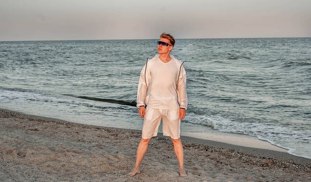 un tipo con ropa blanca mira el atardecer junto al mar - Foto, Imagen