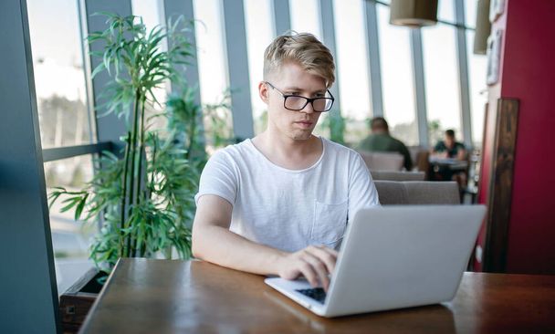 Un joven con una camiseta blanca y gafas trabaja en un portátil sentado en una mesa en una cafetería - Foto, Imagen