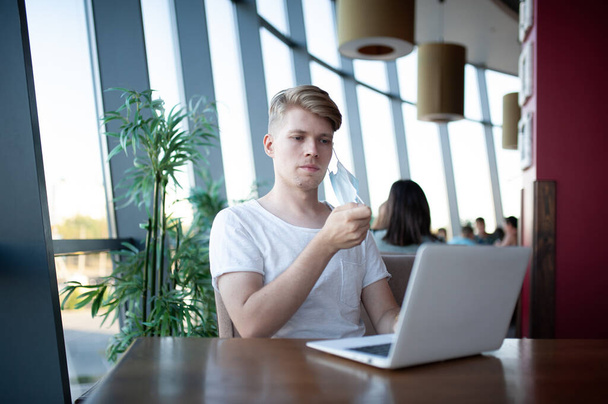 pohledný mladý muž v masce, pracuje na notebooku v kavárně  - Fotografie, Obrázek