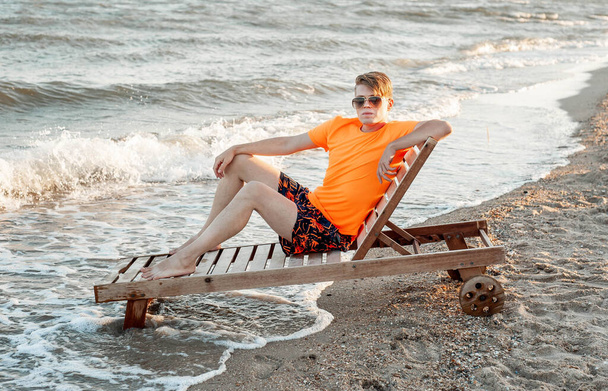 Un ragazzo in maglietta e pantaloncini seduti su una sedia a sdraio in legno vicino al mare - Foto, immagini