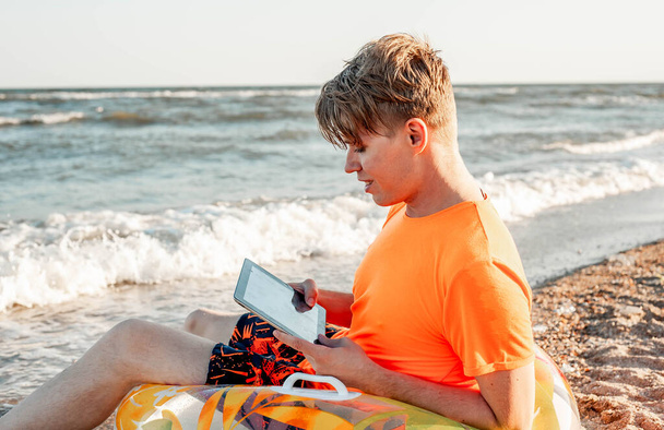 Un mec en T-shirt et short tient une tablette, assis sur une chaise longue en bois au bord de la mer - Photo, image