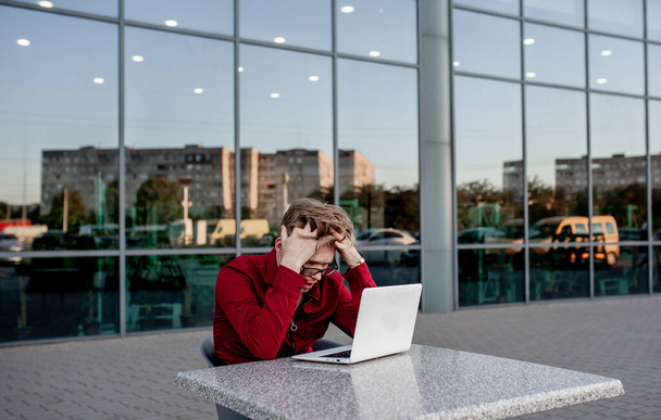 Een jonge man zit aan een tafel op straat en werkt op een laptop. - Foto, afbeelding