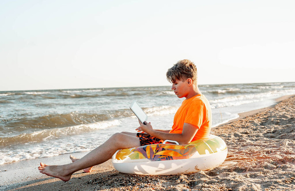 Ein Typ in T-Shirt und kurzer Hose hält Tablet in der Hand, sitzt auf einem hölzernen Liegestuhl am Meer - Foto, Bild