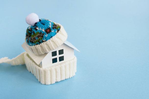 Modèle de maison portant un chapeau et une écharpe tricotés sur une table bleu clair. Système de chauffage, concept hiver ou saison froide. Espace de copie, gros plan - Photo, image