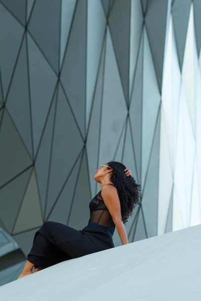 A vertical shot of a curly brunette Hispanic female an elegant black dress sitting under the wall of a modern building with geometric patterns - Φωτογραφία, εικόνα