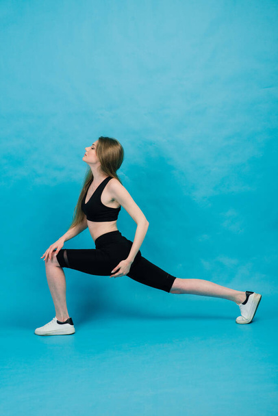 Фитнес. Женщина в спортивной одежде растягивает ноги, согреваясь на синем фоне. - Фото, изображение