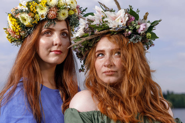 Two young and lovely girls in flower wreaths in nature. Ancient pagan origin celebration concept. Summer solstice day. Mid summer. - Foto, immagini