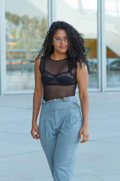 A vertical shot of a curly brunette Hispanic female posing in light blue trousers and a transparent black blouse - Foto, Imagen