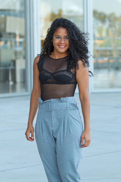 A vertical shot of a curly brunette Hispanic female posing in light blue trousers and a transparent black blouse - Photo, Image