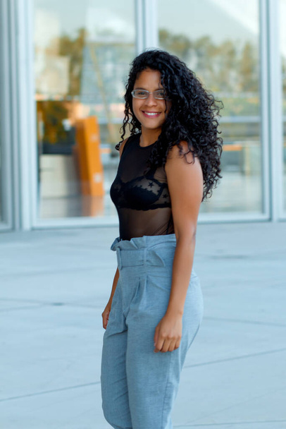 A vertical shot of a curly brunette Hispanic female posing in light blue trousers and a transparent black blouse - Valokuva, kuva
