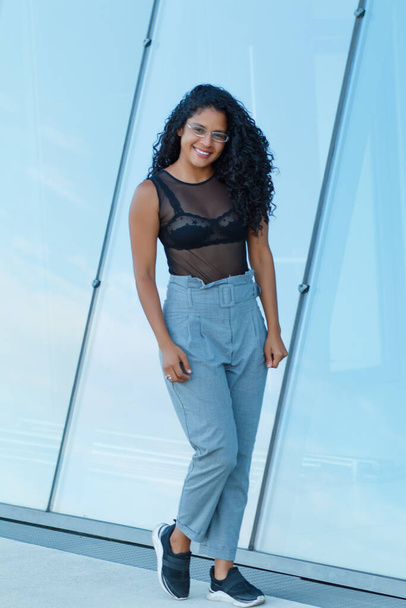 A vertical shot of a curly brunette Hispanic female posing in light blue trousers and a transparent black blouse - Foto, Bild