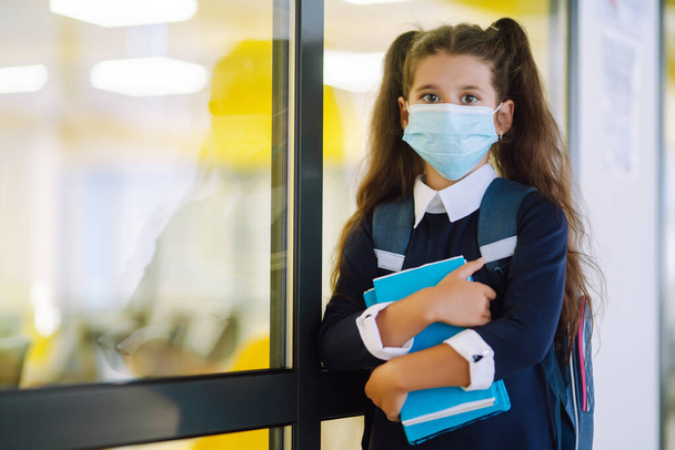 Schoolgirl in a protective mask with a backpack and a textbook in her hands. Security concept, virus protection. Covid-2019. - 写真・画像