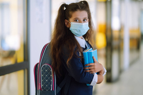 Schoolgirl in a protective mask with a backpack and a textbook in her hands. Security concept, virus protection. Covid-2019. - 写真・画像
