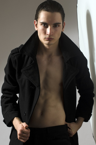 Young man wearing a long black coat and showing his abs and chest muscles, skinny, anorexic latin model - Fotografie, Obrázek