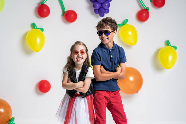 Cool very stylish children in sunglasses and fashionable clothes, posing at background fruit balls. boy and girl stand with their backs to each other and show language concept summer, vitamins, fruits - Foto, afbeelding