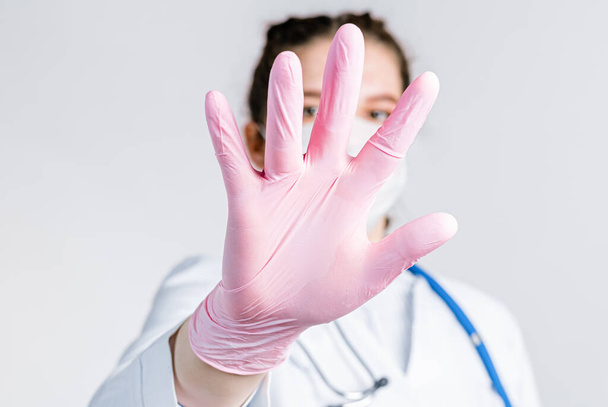 Woman doctor in a white coat and pink sterile gloves close up shows a stop gesture on white background. Say No virus disease. Woman focus on stretched hand as symbol of keep distance avoid communication - Photo, Image