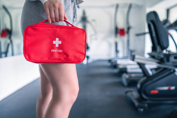 First aid kit red box in the instructor female hand in fitness gym opposite the sport equipment and  jogging simulators. Healthy lifestyle, safety and help concept. - Foto, afbeelding
